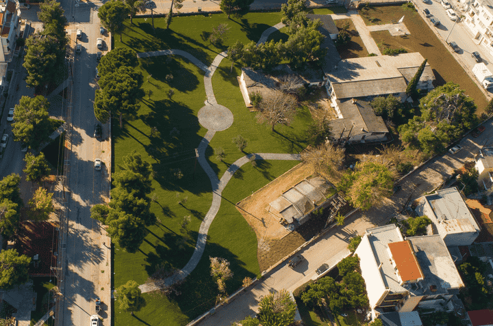 A landscape illustrating a park area with grass, trees, winding pathways, and a central circular paved area, surrounded by buildings and roads.